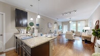 This single-floor residence is designed with an open-concept layout that seamlessly connects the kitchen, dining, and living areas, creating a cohesive and functional living environment. The kitchen is outfitted with dark espresso cabinetry, stainless steel appliances, and a granite-topped peninsula with bar seating, illuminated by pendant lighting and a track light system overhead. Crown molding adds a refined architectural detail throughout the main living spaces.

The living room is generously proportioned, featuring rich hardwood-style flooring, crown molding, and a ceiling fan that enhances natural light drawn in through large windows overlooking greenery outside. An arched passageway connects the living area to the private quarters of the home.

The bedroom is carpeted for comfort and receives natural light through a window dressed with sheer curtains and blinds. It is complemented by a spacious walk-in closet with wraparound wire shelving, offering substantial storage capacity. The full bathroom features wood-look vinyl flooring, a dark-cabineted vanity with a sink, and a full tub-shower combination with white tile surround. Neutral warm tones carry throughout, contributing to a calm and cohesive ambiance.