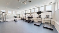 This fitness center offers a comprehensive, professionally designed exercise facility spread across a single floor. The space is divided into two distinct zones: a main workout area finished with durable rubber flooring, and an adjoining studio area featuring light hardwood floors, soaring ceilings, and an elegant chandelier that adds a refined touch to the space.

The main exercise room is bright and airy, with a row of clerestory windows along the upper walls that flood the interior with natural light. The room is equipped with a full lineup of cardio machines, including treadmills, ellipticals, and a stationary bike, many paired with individual screens for entertainment. A mirrored wall expands the sense of space, while wall-mounted flat-screen TVs provide additional viewing options. A dumbbell rack and multi-station cable machine round out the strength training offerings. Recessed lighting and crisp white walls contribute to a clean, motivating atmosphere.

The adjacent studio area features large sliding glass doors that connect the two zones and offer views through to restroom facilities. A rowing machine and spin bike anchor the studio space, which is ideal for low-impact or flexible training. Built-in cubbies along the wall provide convenient storage for personal items, making this fitness center as functional as it is well-appointed.