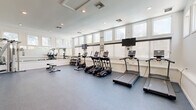 This fitness center offers a comprehensive, professionally designed exercise facility spread across a single floor. The space is divided into two distinct zones: a main workout area finished with durable rubber flooring, and an adjoining studio area featuring light hardwood floors, soaring ceilings, and an elegant chandelier that adds a refined touch to the space.

The main exercise room is bright and airy, with a row of clerestory windows along the upper walls that flood the interior with natural light. The room is equipped with a full lineup of cardio machines, including treadmills, ellipticals, and a stationary bike, many paired with individual screens for entertainment. A mirrored wall expands the sense of space, while wall-mounted flat-screen TVs provide additional viewing options. A dumbbell rack and multi-station cable machine round out the strength training offerings. Recessed lighting and crisp white walls contribute to a clean, motivating atmosphere.

The adjacent studio area features large sliding glass doors that connect the two zones and offer views through to restroom facilities. A rowing machine and spin bike anchor the studio space, which is ideal for low-impact or flexible training. Built-in cubbies along the wall provide convenient storage for personal items, making this fitness center as functional as it is well-appointed.
