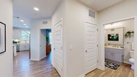 This single-floor residence is thoughtfully designed with a clean, contemporary aesthetic defined by crisp white walls, wood-look flooring, and an abundance of natural light. The open-concept kitchen and living area serves as the heart of the home, where a well-appointed kitchen features white shaker cabinetry, stainless steel appliances, quartz countertops, and a central island with seating for casual dining. Pendant lighting with metallic gold accents adds a stylish focal point overhead. The adjacent living area is generously proportioned, benefiting from a striking bank of large windows that flood the space with daylight and offer outdoor views, creating a bright and welcoming atmosphere. A wide, light-filled hallway connects the main living areas to the private rooms, which include multiple bedrooms. Each bedroom is comfortably sized with windows that bring in natural light. The primary suite includes access to a walk-in closet outfitted with wire shelving systems, providing ample storage. The bathroom features a tiled soaking tub and shower combination. Additional closet storage is accessible throughout the unit. Recessed lighting and ceiling-mounted HVAC vents contribute to the polished, move-in-ready finish of this well-designed home.