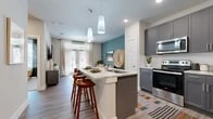 This single-story residence is thoughtfully designed with a contemporary aesthetic that blends functionality and style. The open-concept layout connects the kitchen, dining, and living areas into one cohesive space, creating an effortless flow throughout.

The kitchen is a standout feature, showcasing gray shaker-style cabinetry, light granite countertops, a full suite of stainless steel appliances, and a white subway tile backsplash. A central island with seating provides additional prep space and a casual dining option, illuminated by sleek pendant lighting overhead. Wood-look flooring runs throughout the main living areas, adding warmth and continuity.

The living area is anchored by a striking teal accent wall, French doors that invite natural light, and a ceiling fan overhead. The space is comfortably proportioned for both relaxing and entertaining.

The bedroom is a calm retreat with plush carpeting, a large window with outdoor views, and access to a generously sized walk-in closet fitted with dual hanging rods and built-in shelving for ample storage.

The bathroom features dark cabinetry paired with granite countertops, a wide vanity mirror, wood-look tile flooring, and a glass-enclosed walk-in shower, completing the refined and cohesive design of the space.