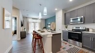This single-story residence is thoughtfully designed with a contemporary aesthetic that blends functionality and style. The open-concept layout connects the kitchen, dining, and living areas into one cohesive space, creating an effortless flow throughout.

The kitchen is a standout feature, showcasing gray shaker-style cabinetry, light granite countertops, a full suite of stainless steel appliances, and a white subway tile backsplash. A central island with seating provides additional prep space and a casual dining option, illuminated by sleek pendant lighting overhead. Wood-look flooring runs throughout the main living areas, adding warmth and continuity.

The living area is anchored by a striking teal accent wall, French doors that invite natural light, and a ceiling fan overhead. The space is comfortably proportioned for both relaxing and entertaining.

The bedroom is a calm retreat with plush carpeting, a large window with outdoor views, and access to a generously sized walk-in closet fitted with dual hanging rods and built-in shelving for ample storage.

The bathroom features dark cabinetry paired with granite countertops, a wide vanity mirror, wood-look tile flooring, and a glass-enclosed walk-in shower, completing the refined and cohesive design of the space.