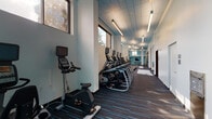This fitness center is designed to support a comprehensive workout experience across a thoughtfully organized single-floor layout. The space is divided into distinct functional zones, each catering to different training needs. A dedicated cardio corridor runs along a window-lined wall, housing a row of elliptical trainers, stationary bikes, and treadmills from Matrix and similar commercial brands, all benefiting from natural light and outdoor views that create an open, energizing atmosphere.

Adjacent to the cardio area, a strength and functional training zone features a multi-cable machine, free weights, resistance equipment, and a rowing machine. This section is enclosed by floor-to-ceiling glass panels, providing visual openness while defining the space. A separate area accommodates flexibility and functional training, equipped with stability balls, medicine balls, resistance bands, and a wall-mounted television for guided workouts. Built-in shelving units and storage racks keep accessories organized throughout.

The interior design maintains a clean, modern aesthetic with teal accent walls, white structural columns, drop tile ceilings with linear pendant lighting, and dark striped carpeting with blue detailing. Large windows throughout admit generous natural light, contributing to an energetic yet focused training environment.