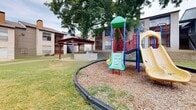 Located at Park Springs Circle in Arlington, Texas, this outdoor community space serves as a central recreational area within a multi-building residential complex. The grounds are well-maintained, featuring open green lawns that provide ample room for outdoor activities and leisure.

At the heart of the space is a colorful multi-component playground structure set within a wood chip-lined play area bordered by a low edging rail. The play set includes a yellow slide, climbing elements, and a tower, offering a variety of recreational options. Mature trees shade portions of the yard, creating a comfortable outdoor environment.

Adjacent to the playground is a wooden pergola structure that provides a shaded gathering spot, accompanied by an outdoor seating area with a table and chairs. A raised stone retaining wall runs along one side of the property, adding a sturdy and attractive landscape feature that separates elevation changes across the grounds.

The surrounding buildings feature beige horizontal lap siding with balconies and exterior staircases, contributing to the cohesive aesthetic of the community. A central air conditioning unit and pathway are also visible along the exterior. The overall atmosphere is a functional, community-oriented outdoor space designed to support both active play and relaxed outdoor enjoyment for residents.