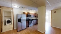 This single-floor space includes a well-appointed kitchen that serves as a central feature of the layout. The kitchen is outfitted with a full suite of stainless steel appliances, including a side-by-side refrigerator with an in-door ice and water dispenser, a freestanding range, and an over-the-range microwave. Dark granite or granite-style countertops provide a sleek contrast against the warm-toned, medium-brown cabinetry that lines the walls, offering generous storage space. A track lighting system mounted on the ceiling directs focused illumination across the workspace, complementing the flush-mount ceiling fixtures overhead. The flooring throughout is light-toned hardwood, lending warmth and continuity to the space. A structural column near the kitchen entrance defines the boundary between the kitchen and an adjacent area, which appears to benefit from natural light through large windows visible in the background. The drop ceiling with grid-style tiles gives the space a clean, functional character. Overall, the space presents a practical and organized environment with a cohesive design palette of warm wood tones and stainless steel accents.