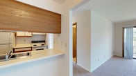 This single-floor space is designed with a functional and straightforward layout, incorporating a mix of flooring materials and practical finishes throughout. The kitchen is configured as a galley-style space, featuring white cabinetry with warm wood trim accents, light countertops, and white appliances including a refrigerator, electric range, and dishwasher. A double basin sink and tile-style flooring complete the kitchen's utilitarian design. A pass-through counter connects the kitchen to the adjacent living area, enhanced by a wood-framed overhead cabinet that adds a natural element to the open sightline.

The living area transitions between plush carpeting and hardwood-style flooring near the entryway, where a solid wood door provides a welcoming focal point. Carpeted bedrooms offer a comfortable feel underfoot, with at least one bedroom featuring a window that allows natural light and views of a green outdoor setting.

The bathroom is well-appointed with a dual-sink vanity topped with a continuous white countertop, white cabinetry below, a large wall mirror, and a full tub-shower combination. Light tile flooring and bright overhead lighting give the bathroom a clean, bright appearance. Throughout the space, neutral white walls and simple finishes create a consistent, move-in-ready aesthetic.