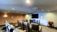 This single-floor theater room is thoughtfully designed to deliver a premium entertainment experience. The space is anchored by a large flat-screen television mounted on a striking geometric-patterned accent wall, complemented by a dark wood media console that houses audio-visual equipment. Rows of plush, dark leather recliner chairs are arranged in a tiered layout, ensuring clear sightlines from every seat and providing a comfortable, cinema-style atmosphere. Recessed lighting is embedded throughout the ceiling, offering adjustable illumination that can be dimmed to set the mood for movie viewing. The walls feature a combination of warm neutral tones and a bold geometric wallpaper panel, adding visual depth and character to the room. Carpeted flooring in a subtle pattern contributes to the acoustic quality of the space while enhancing its overall aesthetic. A small decorative floral arrangement adds a welcoming touch to the room. The thoughtful design and quality furnishings make this theater space well-suited for group screenings, entertainment events, or relaxed viewing sessions.