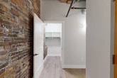 Located at 119 Hotel St in Utica, NY, this 2-bedroom, 2-bathroom unit blends industrial character with contemporary design. The open-concept living and kitchen area is anchored by soaring ceilings with exposed wooden beams and visible ductwork, complemented by light hardwood flooring throughout. The kitchen is a standout feature, outfitted with two-tone cabinetry — white upper cabinets and gray lower cabinets — paired with quartz countertops, a subway tile backsplash, stainless steel appliances, and a central island with pendant lighting above. Exposed brick walls carry through the living areas and bedrooms, adding warmth and historic texture. The primary bedroom is generously sized, featuring exposed brick, large windows, and original wooden beam accents. Three closets provide ample storage, including a walk-in closet that continues the exposed brick aesthetic. Both bathrooms are finished to a high standard, with hexagonal mosaic tile flooring, dark navy vanities, quartz countertops, and backlit mirrors. The primary bath features a double vanity and glass-enclosed shower, while the second bath includes a tub-shower combination. A connecting hallway ties the layout together efficiently, creating a cohesive and well-organized living space with strong industrial-modern appeal.