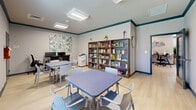 Located at 2001 Amber Leaf Pl in Waldorf, Maryland, this single-room office is a thoughtfully designed, multipurpose space that balances productivity and collaboration. Light hardwood flooring runs throughout, complementing the neutral gray walls accented by a deep teal crown molding that adds a polished, distinctive touch to the room's aesthetic.

The space is divided into functional zones. Along one wall, a long wooden desk supports multiple computer workstations with ergonomic task chairs, accompanied by a multifunction printer — making it well-suited for administrative tasks and document management. Adjacent to the workstation area, a tall double-unit bookshelf houses a broad collection of books and resources, adding both utility and visual warmth to the room.

The collaborative seating area features multiple square blue-topped tables paired with modern light gray chairs, providing ample space for group work, meetings, or study sessions. Natural light filters in through windows fitted with horizontal blinds, offering views of the surrounding greenery outside. Flush-mounted ceiling light panels ensure consistent, bright illumination throughout.

Decorative wall art, including vibrant floral panels and a landscape painting, contribute to a welcoming and engaging atmosphere. The room is equipped with standard safety features, including a fire extinguisher, and includes a secondary door for convenient access.