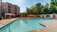 Located at 1315 Creekshire Way in Winston-Salem, NC, this outdoor amenity area is a standout communal space designed for relaxation and recreation. At its centerpiece is a generously sized, resort-style swimming pool with clear blue water, gradual depth transitions, and multiple entry points via stainless steel railings. A separate hot tub offers a quieter retreat adjacent to the main pool. The pool deck is finished in a smooth concrete surround and is furnished with rows of chaise lounge chairs, providing ample space for sunbathing and leisure. A charming stone-accented gazebo with a pitched roof serves as a shaded gathering spot, while multiple white pergolas add architectural character and additional shade throughout the deck. The space is enclosed by a classic brick perimeter wall, enhancing the sense of privacy and cohesion. A dedicated poolside structure with large windows—likely a clubhouse or amenity building—provides interior access to additional facilities. Mature trees line the backdrop of the space, offering natural shade and a scenic green canopy. The multi-story brick residential building frames the courtyard on multiple sides, creating an intimate, enclosed atmosphere. Overall, the space conveys a well-appointed, community-focused outdoor environment ideal for year-round enjoyment.