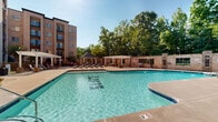 Located at 1315 Creekshire Way in Winston-Salem, NC, this outdoor amenity area is a standout communal space designed for relaxation and recreation. At its centerpiece is a generously sized, resort-style swimming pool with clear blue water, gradual depth transitions, and multiple entry points via stainless steel railings. A separate hot tub offers a quieter retreat adjacent to the main pool. The pool deck is finished in a smooth concrete surround and is furnished with rows of chaise lounge chairs, providing ample space for sunbathing and leisure. A charming stone-accented gazebo with a pitched roof serves as a shaded gathering spot, while multiple white pergolas add architectural character and additional shade throughout the deck. The space is enclosed by a classic brick perimeter wall, enhancing the sense of privacy and cohesion. A dedicated poolside structure with large windows—likely a clubhouse or amenity building—provides interior access to additional facilities. Mature trees line the backdrop of the space, offering natural shade and a scenic green canopy. The multi-story brick residential building frames the courtyard on multiple sides, creating an intimate, enclosed atmosphere. Overall, the space conveys a well-appointed, community-focused outdoor environment ideal for year-round enjoyment.