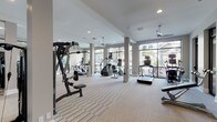 This well-appointed fitness center offers a thoughtfully designed layout across a single level, providing ample space for a full range of workout activities. The main exercise area is defined by neutral-toned carpet flooring with a subtle stripe pattern, structural columns, and ceiling fans that contribute to a comfortable environment during use. Floor-to-ceiling windows along one wall flood the space with natural light while offering views of the surrounding outdoor landscape, creating an open, airy atmosphere.

The cardio zone is equipped with treadmills, stair climbers, elliptical machines, and stationary bikes positioned to take advantage of the natural light and exterior views. A dedicated strength training room, enclosed by sleek black-framed glass walls and doors, houses a robust selection of cable machines, a Smith machine, a power rack, adjustable benches, and free weights. Full-length mirrors line the walls of this area, further enhancing the sense of space and allowing users to monitor their form. Wall-mounted flat-screen televisions are positioned throughout for added convenience. Additional storage shelving and a water station are also present, rounding out the facility's practical amenities.