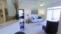 This single-floor residence offers a thoughtfully designed layout that blends comfort and style. The living area serves as the heart of the space, featuring plush wall-to-wall carpeting that adds warmth underfoot, elegant crown molding that frames the room with a refined architectural detail, and a ceiling fan that provides both function and a classic finishing touch. Natural light filters in through window treatments, lending the space a bright and airy ambiance. The living room is generously proportioned, with ample room for a sofa, accent chairs, and a glass coffee table, creating a comfortable and inviting gathering area. A decorative wall-mounted artwork adds a splash of color above the seating arrangement, complemented by coordinating accent pillows in rich blue tones. A counter or bar-height surface transitions from an adjacent area into the living space, hinting at an open-concept flow. A doorway leads to a connected bedroom, visible in the background, which features its own window and dedicated lighting. The overall ambiance is clean, neutral, and modern, with a soft color palette of whites, grays, and blues that creates a calm and welcoming atmosphere throughout.