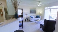 This single-floor residence offers a thoughtfully designed layout that blends comfort and style. The living area serves as the heart of the space, featuring plush wall-to-wall carpeting that adds warmth underfoot, elegant crown molding that frames the room with a refined architectural detail, and a ceiling fan that provides both function and a classic finishing touch. Natural light filters in through window treatments, lending the space a bright and airy ambiance. The living room is generously proportioned, with ample room for a sofa, accent chairs, and a glass coffee table, creating a comfortable and inviting gathering area. A decorative wall-mounted artwork adds a splash of color above the seating arrangement, complemented by coordinating accent pillows in rich blue tones. A counter or bar-height surface transitions from an adjacent area into the living space, hinting at an open-concept flow. A doorway leads to a connected bedroom, visible in the background, which features its own window and dedicated lighting. The overall ambiance is clean, neutral, and modern, with a soft color palette of whites, grays, and blues that creates a calm and welcoming atmosphere throughout.