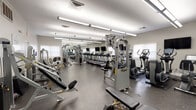 This fitness center is a comprehensively outfitted single-floor facility designed to support a full range of workout routines. The space is finished with durable rubber flooring throughout and illuminated by a series of linear fluorescent light fixtures that provide consistent, even lighting across the entire area. Large wall-mounted mirrors run along multiple sides of the room, enhancing the sense of openness and allowing users to monitor their form during exercise.

The cardio zone features a row of treadmills and elliptical machines, each equipped with individual digital consoles and paired with wall-mounted flat-screen televisions for entertainment during workouts. Stationary upright bikes are also available in this section. Natural light filters in through several windows fitted with white blinds, contributing to the bright and functional atmosphere.

The strength training area includes a selection of Hoist-brand cable and selectorized machines, accommodating a variety of muscle groups. A dedicated free weights section offers a full rack of dumbbells and a tiered kettlebell rack, accompanied by adjustable benches. French doors along one wall provide access to an adjacent area, while large mirrors further visually expand the space. The overall layout is organized, clean, and practical.
