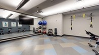 This fitness room is thoughtfully designed to accommodate a wide range of exercise activities. The space features a clean, open layout with a patterned tile floor in soft blue, beige, and gray tones, providing a durable and easy-to-maintain workout surface. A large mirrored wall spans one side of the room, enhancing the sense of space while allowing users to monitor their form during exercise. The room is outfitted with a variety of equipment, including multiple dumbbell racks with graduated weights, a collection of stability balls in assorted sizes, and a TRX suspension training system mounted to the wall. A ceiling fan ensures comfortable air circulation throughout the space. Recessed and track lighting provide bright, even illumination across the entire room. The neutral-toned walls and organized equipment layout give the space a clean, professional atmosphere. The open floor area allows for stretching, bodyweight exercises, and functional fitness movements. Overall, this fitness room is a practical and versatile space well-suited for both individual strength training and group workout sessions.
