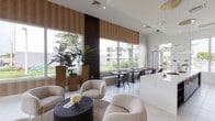 This elegantly designed single-floor amenity space presents a seamless blend of modern aesthetics and functional comfort. The open-plan layout connects a generous lounge area, a casual dining zone, and a fully equipped kitchen, creating an inviting environment suited for both social gatherings and relaxed lounging.

The lounge area is anchored by plush, cream-toned curved armchairs and deep navy sectional sofas with gold-accented legs, arranged around dark wood coffee tables. Soaring double-height ceilings amplify the sense of space, while floor-to-ceiling sheer curtains frame oversized windows that flood the interior with natural light and offer panoramic views of the surrounding greenery and palm trees.

Textured warm-toned wall panels add depth and contrast against the crisp white tile flooring, which runs throughout the space. Tall architectural planters with tropical foliage reinforce the connection between indoors and outdoors.

The kitchen is a standout feature, showcasing an expansive white quartz island with navy velvet bar seating trimmed in gold. Rich espresso-toned cabinetry lines the wall, complemented by open display shelving and dramatic globe pendant lighting. Stainless steel appliances complete the professional-grade setup. Together, these elements create a polished, contemporary atmosphere that is both visually striking and highly functional.