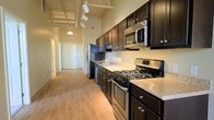 This single-floor space showcases an open, elongated layout with a well-appointed galley-style kitchen as a central feature. The kitchen is outfitted with rich, dark espresso cabinetry — both upper and lower — providing ample storage, complemented by light granite countertops that create a striking contrast. Stainless steel appliances, including a gas range, built-in microwave, and refrigerator, lend a modern, functional character to the space.

The flooring throughout is light hardwood, adding warmth and continuity as it extends from the kitchen into the long hallway beyond. Notable above are the tall ceilings fitted with exposed industrial-style track lighting, adding both visual height and practical illumination to the kitchen area. A pendant light further down the hallway contributes to the layered lighting scheme.

The hallway stretches toward additional rooms, suggesting a thoughtful linear floor plan. White interior doors and neutral wall tones keep the space feeling bright and clean. The overall ambiance balances industrial elements with warm, contemporary finishes, creating a practical yet stylish living environment.