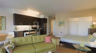 This well-designed single-floor studio offers a thoughtfully organized open-concept layout that brings together the living, sleeping, and kitchen zones in one cohesive space. The kitchen is outfitted with dark espresso cabinetry that extends to the ceiling, providing ample storage while creating a bold visual contrast against the light-toned walls. Stainless steel appliances, including a refrigerator, range, and built-in microwave, lend a sleek, modern feel, while the under-cabinet lighting brightens the workspace. A compact dining nook sits adjacent to the kitchen, making efficient use of the available floor plan. The living area features warm hardwood-style flooring that flows throughout the entire space, anchoring a comfortable seating arrangement accented with a vibrant green sofa and colorful throw pillows. A piece of wall art adds a personal, welcoming touch to the neutral-toned walls. The sleeping area is integrated seamlessly into the open floor plan, offering a practical and unobstructed layout. The smooth, white ceilings and clean architectural lines contribute to an overall sense of brightness and openness, making this compact space feel both functional and inviting.