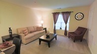This single-floor space features a thoughtfully designed living room that serves as its central gathering area. The room is finished with wall-to-wall carpeting in a neutral beige tone, contributing to a warm and comfortable atmosphere. Soft yellow-toned walls enhance the sense of warmth throughout the space.

A large sofa anchors the seating area, complemented by an accent chair upholstered in deep burgundy, adding a pop of color against the neutral backdrop. A dark-finished rectangular coffee table sits at the center, providing both function and visual contrast. A side table with a classic table lamp offers additional ambient lighting alongside the room's overhead fixtures.

Natural light enters through a double window dressed with decorative patterned curtains in rich burgundy and brown tones, framing the window attractively while allowing daylight to filter through. The window provides a connection to the outdoors and contributes to the bright, airy feel of the room.

The overall ambiance is cozy and inviting, with a cohesive color palette of warm neutrals accented by deeper jewel tones, creating a balanced and comfortable living environment.