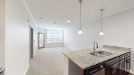 This single-story residence at 13957 College Blvd in Olathe, Kansas, offers a well-designed open-concept layout that seamlessly connects the kitchen and living areas. The kitchen serves as a central hub, showcasing granite countertops atop dark espresso cabinetry, a large island with a stainless steel sink and pendant lighting overhead, a tile backsplash, and a full suite of stainless steel appliances including a refrigerator, range, microwave, and dishwasher. Rich hardwood-style flooring grounds the kitchen and entry areas, adding warmth to the clean, neutral palette throughout.

The living area is generously proportioned, featuring plush carpeting, crown molding, and a wall of windows that invite abundant natural light. A glass door provides direct outdoor access. The bedroom continues the carpeted comfort, accented by crown molding, a ceiling fan, and a bright window. It connects to a walk-in closet with built-in shelving and a full bathroom featuring granite countertops on dark cabinetry, a framed mirror, and a tub-shower combination set on hardwood-style flooring. The overall ambiance is clean, modern, and move-in ready.