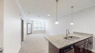 This single-story residence at 13957 College Blvd in Olathe, Kansas, offers a well-designed open-concept layout that seamlessly connects the kitchen and living areas. The kitchen serves as a central hub, showcasing granite countertops atop dark espresso cabinetry, a large island with a stainless steel sink and pendant lighting overhead, a tile backsplash, and a full suite of stainless steel appliances including a refrigerator, range, microwave, and dishwasher. Rich hardwood-style flooring grounds the kitchen and entry areas, adding warmth to the clean, neutral palette throughout.

The living area is generously proportioned, featuring plush carpeting, crown molding, and a wall of windows that invite abundant natural light. A glass door provides direct outdoor access. The bedroom continues the carpeted comfort, accented by crown molding, a ceiling fan, and a bright window. It connects to a walk-in closet with built-in shelving and a full bathroom featuring granite countertops on dark cabinetry, a framed mirror, and a tub-shower combination set on hardwood-style flooring. The overall ambiance is clean, modern, and move-in ready.