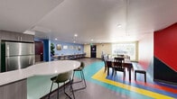Located at 3515 Vista Ln in San Diego, CA, this single-floor space offers a thoughtfully designed layout that blends functionality with a lively, welcoming atmosphere. The interior is immediately distinguished by its bold use of color — vivid multicolored flooring strips in red, yellow, green, and blue run throughout the common areas, contrasting against soft sage-green and warm neutral wall tones punctuated by a striking red accent wall.

The living area is generously proportioned, furnished with rich brown leather reclining sofas that invite relaxation. A large circular area rug and recessed lighting add warmth and definition to the open floor plan. The space transitions naturally into a flexible activity or lounge zone, complete with seating arrangements and a dining table with chairs — ideal for group gatherings.

The kitchen is modern and well-appointed, featuring charcoal-toned cabinetry, a stainless steel refrigerator, and a sleek white oval island with stylish green upholstered bar stools. Light mint-green walls and polished countertops give the kitchen a clean, contemporary feel.

Throughout the space, recessed ceiling lighting ensures consistent illumination, while vertical blinds on the windows allow for natural light control. The overall ambiance is energetic yet comfortable, making this a versatile space well-suited for community engagement, programming, or shared use.