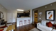 This single-floor residence offers a thoughtfully designed open-concept layout that seamlessly connects the living and kitchen areas into one cohesive, functional space. Rich, dark hardwood flooring runs throughout, grounding the space with warmth and a sense of continuity.

The kitchen is outfitted with dark cabinetry that extends to the upper walls, providing ample storage, and is equipped with a built-in microwave and additional appliances. A white breakfast bar peninsula serves as the centerpiece dividing the kitchen from the living area, accompanied by three copper-toned metal barstools that add a touch of industrial character. The countertop space is generous, offering plenty of room for meal preparation.

The living area is bright and welcoming, featuring a neutral-toned sofa with patterned accent pillows, a area rug, and a coffee table — creating a comfortable and inviting atmosphere. An accent wall in a deep taupe tone adds visual depth and contrast to the otherwise light-colored walls.

Just off the main living space, a dedicated laundry area is visible, housing a full-size washer and dryer — a convenient and practical feature. The front entryway is framed by a classic white paneled door, completing the clean, cohesive aesthetic of this well-appointed residence.