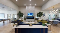 This single-floor community clubhouse is thoughtfully designed to offer a variety of social and recreational spaces, all unified by a cohesive modern aesthetic. The welcoming lobby greets visitors with herringbone hardwood flooring, bold botanical-print drapery, an eye-catching sculptural gold chandelier, and workstation desks accented by blue chairs — creating a polished yet approachable environment. Arched windows flood the space with natural light, while a wall-mounted display and seating areas provide functional comfort.

The central lounge is a standout feature, anchored by a dramatic floor-to-ceiling patterned tile accent wall housing a sleek electric fireplace and mounted television. Plush navy and olive-green velvet sofas and armchairs are arranged for conversation, complemented by lush fiddle-leaf fig trees that bring a touch of nature indoors. Light wood herringbone floors carry throughout, adding warmth and continuity.

Adjacent areas include a casual dining space with a round marble-top table, patterned upholstered chairs, and a stylish bubble chandelier overhead, alongside navy cabinetry and a beverage bar. A game room with a full-size billiards table and additional lounge seating offers recreational appeal. A window bar counter with gold metal stools overlooks a lushly landscaped outdoor courtyard with arched architectural details, providing a bright and inviting spot to relax.