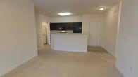 This single-floor unit offers an open-concept design that integrates the kitchen and living area into one cohesive space. The kitchen is defined by dark espresso-toned cabinetry that contrasts with the light neutral walls, and it is equipped with black appliances, including a refrigerator and microwave. A white peninsula counter extends outward, providing additional workspace and serving as a natural divider between the kitchen and the living area. The flooring transitions from tile in the kitchen and entryway zone to wall-to-wall carpet in the living area, adding warmth and comfort underfoot. The layout is notably open, allowing for flexible furniture arrangements within the living space. A doorway near the kitchen leads to a laundry area, where a washer and dryer are visible, adding practical convenience. An exterior door is positioned adjacent to the kitchen, offering a secondary point of entry. The unit is illuminated by flush-mount ceiling lights, maintaining a clean and uncluttered aesthetic throughout. Neutral tones across the walls, flooring, and trim create a cohesive, move-in-ready environment.