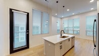 Located at 6505 Rockhill Rd in Kansas City, Missouri, this modern single-floor unit showcases a clean, contemporary design throughout. The open-concept kitchen is a standout feature, centered around a large white island with an integrated sink and matte black fixtures, elegant pendant lighting, and stainless steel appliances including a gas range, built-in microwave, and French door refrigerator. A bold navy tile backsplash adds a striking contrast to the white cabinetry. Light hardwood flooring flows through the main living and kitchen areas, while abundant natural light pours in through multiple large windows fitted with horizontal blinds. The living area extends seamlessly from the kitchen, enhanced by recessed lighting and access to an exterior door. The bedrooms feature plush carpeting, ceiling fans, generous windows with exterior views, and sleek black sliding closet doors that contribute to the modern aesthetic. Two bathrooms serve the unit — one features a white vanity with a clean countertop and a shower-tub combination, while the second is a more spacious, accessibility-equipped bathroom with grab bars, a roll-in shower, and hardwood-style flooring. The overall ambiance is bright, clean, and move-in ready, with a consistent modern design palette throughout.
