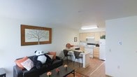 This single-floor space showcases an open-concept design that integrates the living area and kitchen into one cohesive, functional layout. The living area features smooth white walls and light wood-style flooring that extends throughout, creating a warm and unified aesthetic. A large framed wall art piece adds a decorative focal point, while the space accommodates comfortable seating alongside a compact dining area with a small table and chairs — ideal for casual meals.

The kitchen is situated directly adjacent to the living space, benefiting from a flush transition that enhances the sense of openness. It is equipped with light wood cabinetry, white appliances including a refrigerator, oven range, and microwave, as well as a functional countertop workspace. Overhead fluorescent lighting brightens the kitchen area effectively. A peninsula-style counter separates the kitchen from the dining nook, offering additional surface space and a natural visual boundary between zones.

The overall ambiance is clean, bright, and practical, with neutral tones throughout that create a welcoming atmosphere. The thoughtful layout maximizes the available space while maintaining clear, open sightlines across the unit.