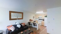 This single-floor space showcases an open-concept design that integrates the living area and kitchen into one cohesive, functional layout. The living area features smooth white walls and light wood-style flooring that extends throughout, creating a warm and unified aesthetic. A large framed wall art piece adds a decorative focal point, while the space accommodates comfortable seating alongside a compact dining area with a small table and chairs — ideal for casual meals.

The kitchen is situated directly adjacent to the living space, benefiting from a flush transition that enhances the sense of openness. It is equipped with light wood cabinetry, white appliances including a refrigerator, oven range, and microwave, as well as a functional countertop workspace. Overhead fluorescent lighting brightens the kitchen area effectively. A peninsula-style counter separates the kitchen from the dining nook, offering additional surface space and a natural visual boundary between zones.

The overall ambiance is clean, bright, and practical, with neutral tones throughout that create a welcoming atmosphere. The thoughtful layout maximizes the available space while maintaining clear, open sightlines across the unit.