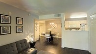 This single-floor residence offers a thoughtfully designed open-concept layout that connects the living, dining, and kitchen areas into one cohesive and functional living space. Rich, dark hardwood floors run throughout, providing a warm and elegant foundation that complements the light, neutral-toned walls.

The living area features a contemporary grey leather sofa paired with a sleek side table and a decorative lattice lamp, creating a comfortable and stylish retreat. Natural light filters in, casting soft patterns across the walls and adding to the inviting ambiance.

The dining area is defined by a bold black rectangular table surrounded by modern white chairs, anchored beneath a geometric chandelier that serves as a striking focal point. A decorative wood-panel wall accent adds texture and visual interest to the space.

Adjacent to the dining area, the kitchen is bright and clean, featuring white cabinetry, white appliances, and light countertops, offering a crisp contrast to the darker tones of the living and dining spaces. A passthrough counter connects the kitchen to the dining area, enhancing the open and airy feel of the layout.

The space also includes a hallway leading to additional rooms, with built-in shelving adding both practical storage and character to the interior.