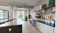 Located at 1775 Ashlar Dr in Fort Collins, Colorado, this single-floor community space offers a thoughtfully designed gathering area with a cohesive modern aesthetic. The open layout flows seamlessly between a fully equipped kitchen, a comfortable living room, and a recreation zone, creating a versatile environment for socializing and relaxation.

The kitchen is outfitted with sleek gray cabinetry, dark countertops, and a patterned geometric tile backsplash that adds visual interest. Floating wood shelves display plants, books, and decorative items, while stainless steel appliances and a beverage refrigerator provide practical functionality. Light wood-look flooring runs throughout, tying the spaces together.

The living area is a standout, anchored by floor-to-ceiling black-framed windows that flood the room with natural light and offer open views of the surrounding outdoor amenities and greenery. Exposed industrial ductwork adds an urban character to the high ceilings, while sheer white drapery softens the look. Comfortable seating, a bold patterned area rug, and curated artwork contribute to an inviting ambiance.

A recreation area with a full-size pool table adds an entertaining dimension to the space. With direct access to the outdoors and abundant natural light throughout, this community lounge is both functional and welcoming.