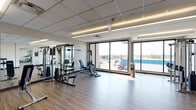 This contemporary fitness facility is thoughtfully designed to support a comprehensive workout experience across a single, open floor. The space is organized into distinct functional zones, each tailored to a specific type of exercise. The main training area features durable wood-look flooring and is lined with large floor-to-ceiling windows that flood the room with natural light and provide open views of the surrounding landscape, creating an energizing atmosphere. This zone is equipped with a multi-station cable machine system, weight benches, and a dumbbell rack positioned in front of full-length mirrors, supporting a wide range of strength training exercises. Adjacent to the strength area, a dedicated cardio section houses treadmills, elliptical trainers, and a rowing machine, all oriented toward expansive windows to enhance the workout experience with outdoor views. A separate, glass-partitioned studio offers a quieter space for stretching, yoga, or mat-based workouts, complete with wall-mounted exercise mats, floor mats, and a mirrored wall. The facility is illuminated throughout by modern recessed linear lighting set within a drop-tile ceiling. Access is provided via a glass-panel entry door, and the overall design reflects a clean, professional aesthetic suited for focused fitness training.