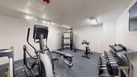 Located at 123 E McCullough Dr in Charlotte, NC, this fitness center is a compact, well-equipped exercise space designed to support a well-rounded workout routine. The room features durable speckled rubber flooring throughout, providing a safe and practical surface for a wide variety of exercises. The walls are finished in a clean white, and the textured ceiling is fitted with a combination of flush fluorescent light fixtures and directional track lighting, ensuring the space is brightly and evenly illuminated.

The facility is outfitted with a solid selection of equipment, including Matrix-brand elliptical trainers, a stationary spin bike, and a multi-functional cable trainer with an attached rack of kettlebells and suspension training accessories. A weight bench is centrally positioned for versatile use alongside the cable system. A three-tier dumbbell rack stocked with a full range of free weights is situated along one wall, complemented by a wall-mounted flat-screen television for entertainment during workouts. A fire sprinkler and smoke detector are also present, underscoring the safety features of the space. Access is provided through a single solid door with a narrow window panel. This fitness center delivers a functional and efficient environment for residents to maintain an active lifestyle.