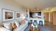 This single-floor residence offers a thoughtfully designed open-concept layout that seamlessly connects the living and kitchen areas, creating an inviting and functional environment. The living room is anchored by a light-toned sofa adorned with decorative pillows, complemented by a plush area rug and a glass-topped coffee table. Built-in floating shelves add a touch of architectural character, providing display space for decorative accents. Warm ambient lighting from a classic table lamp enhances the cozy atmosphere of the space. The kitchen is a standout feature, showcasing dark cabinetry, stainless steel appliances, and a white subway tile backsplash that adds a clean, contemporary contrast. A granite-topped peninsula extends into the dining area, flanked by sleek black metal bar stools, making it an ideal spot for casual dining and entertaining. Pendant lights hang above the peninsula, adding both style and focused illumination. Wood-style flooring runs throughout, tying the open layout together with warmth and continuity. Recessed lighting in the kitchen ensures the space is well-lit and practical. Overall, the residence strikes a balance between modern design and comfortable living, offering a polished yet welcoming ambiance throughout.