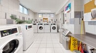 Located at 6151 Bathurst Street in Toronto, this shared laundry facility offers a clean, functional, and well-maintained space designed to serve building residents efficiently. The room features white ceramic floor tiles throughout, providing a bright and easy-to-clean surface underfoot. The walls are finished with a two-tone tile design — white tiles accented by decorative grey mosaic panels — lending the space a tidy, structured appearance.

Multiple front-loading washing machines and dryers are arranged along the perimeter walls, offering ample capacity for simultaneous use. The machines are equipped with digital control panels for ease of operation. Natural light filters in through several windows positioned near the ceiling, complementing the overhead fluorescent lighting that ensures the space remains well-lit at all times.

A folding counter and seating area along one wall provide a convenient spot for sorting and managing laundry. Stainless steel waste bins are positioned near the entrance for cleanliness and convenience. Informational notices and usage guides are posted on the walls, keeping users informed of proper procedures. A decorative potted plant adds a touch of warmth to the otherwise utilitarian environment. The space is thoughtfully organized to maximize usability while maintaining a clean and orderly atmosphere.