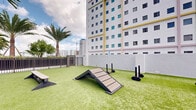 Located at 777 Northwest 2nd Avenue in Miami, FL, this outdoor dog park offers a thoughtfully designed amenity space for pets and their owners in the heart of the city. The area is surfaced with vibrant artificial turf, providing a clean, low-maintenance environment suitable for year-round use in South Florida's climate. The space is fully enclosed by sturdy metal fencing with gated entry and exit points, ensuring a safe, secure area for dogs to roam and play freely. Agility equipment is distributed throughout the turf, including ramps, hurdle bars, and low benches — offering both physical stimulation and training opportunities for dogs of various sizes. Mature palm trees line portions of the perimeter, adding a tropical aesthetic and a sense of greenery against the urban backdrop. The open-air design allows for abundant natural light and open sky views, framing the surrounding Miami skyline and neighboring high-rise buildings. A pergola-style shaded structure provides a comfortable resting spot for owners. The space is well-lit and thoughtfully laid out, balancing functional pet amenities with an inviting, park-like atmosphere that reflects the vibrant character of its Miami location.