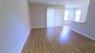 This single-floor space offers a clean, open layout with a warm and inviting ambiance. The main living area is highlighted by rich hardwood-style flooring that runs throughout, reflecting natural light and adding a sense of warmth to the interior. Neutral-toned walls provide a versatile backdrop, complementing the bright and airy atmosphere. A large sliding glass door dressed with vertical blinds serves as a central architectural feature, allowing ample natural light to fill the room while offering access to the outdoors. An additional window further enhances the brightness of the space. The open flow between areas creates a sense of spaciousness, with clean sightlines from the main living area into adjacent spaces. White baseboards provide a crisp contrast against the flooring, adding a polished finishing touch throughout. The overall design is straightforward and functional, offering a move-in ready canvas well-suited to a variety of interior styles. The combination of natural light, consistent flooring, and open layout makes this a comfortable and practical living space.
