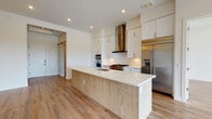 This single-floor space showcases a contemporary design with a cohesive palette of white, warm wood tones, and stainless steel throughout. The kitchen serves as the centerpiece, featuring high-gloss white cabinetry extending to the ceiling for ample storage, a sleek stainless steel range hood, and a professional-grade gas range paired with double wall ovens. A large island with a crisp white quartz countertop and natural wood-paneled base provides generous prep and seating space. A stainless steel French door refrigerator completes the premium appliance suite. Light wood-look flooring flows continuously throughout the space, creating warmth and visual continuity. A sliding barn door adds a functional yet stylish accent near the kitchen entry. The living area benefits from expansive floor-to-ceiling windows and glass sliding doors that open to a balcony, offering panoramic city and treetop views. A tray ceiling with accent lighting adds architectural detail to this room. A dedicated laundry room houses a full-size side-by-side washer and dryer with overhead shelving and a countertop workspace. The bathroom features clean white finishes in a minimalist, private water closet layout. Recessed lighting throughout maintains a bright, modern ambiance.