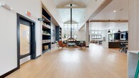 This expansive single-floor community amenity space is defined by its dramatic vaulted ceilings with exposed wood beam detailing and a striking central skylight that floods the interior with natural light. Light-toned hardwood flooring runs throughout, unifying the various zones within the open layout. The main lounge area anchors the space with a central fireplace, a curated seating arrangement of mixed leather and upholstered chairs, and built-in navy blue shelving units that house books, plants, and decorative objects alongside a wall-mounted television. Adjacent to the lounge, a game room offers a billiards table, a large-screen entertainment unit with dark cabinetry, and bold kilim-style area rugs that add warmth and character. A bar counter with vibrant green stools and a patterned tile backdrop provides a casual gathering spot, while intimate booth seating with a tall steel-and-wood shelving display offers a quieter retreat. Black-framed windows and glass doors frame views of an outdoor pool and surrounding community buildings, seamlessly connecting the interior to the outdoors. Decorative wall art and lush indoor plants throughout contribute to a welcoming, modern aesthetic that balances comfort with purposeful design.