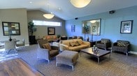 This single-floor space presents a thoughtfully designed lounge area characterized by soaring vaulted ceilings that lend an open, airy quality to the interior. Two large pendant light fixtures hang prominently overhead, casting a warm, inviting glow throughout the room. The ceiling transitions between earthy taupe and cool blue tones, adding visual depth and a modern aesthetic to the space.

The seating arrangement is anchored by a plush camel-toned sofa adorned with decorative pillows, complemented by coordinating upholstered armchairs and an ottoman. A dark wood coffee table serves as a central focal point, styled with tasteful decorative accents. Additional seating groupings are arranged toward the rear of the room, offering versatile options for both intimate conversations and larger gatherings.

Natural light filters in through a horizontal clerestory window along one wall, while a large decorative mirror near the back amplifies the sense of space. Potted tropical plants add organic texture and life to the corners of the room. Carpeted flooring with a subtle pattern grounds the space, while the overall palette of warm neutrals and cool blues creates a calm, contemporary atmosphere throughout.