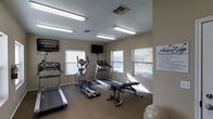 This fitness center is a single-floor space designed to support a well-rounded workout routine. The room is furnished with a selection of cardio equipment, including two treadmills and an elliptical machine, catering to a range of cardiovascular exercise preferences. A weight bench is also available for strength training. Multiple wall-mounted flat-screen televisions are positioned throughout the room, allowing exercisers to stay entertained during their workouts. The space is illuminated by a combination of overhead fluorescent lighting and natural light streaming in through several windows fitted with blinds. The windows offer views of what appears to be an outdoor pool area, adding a pleasant visual connection to the exterior. The floors are covered in a durable, speckled material well-suited for an active environment. The neutral-toned walls contribute to a clean, functional atmosphere. A fitness center guidelines sign is posted on one wall, providing users with the rules and expectations for use of the facility. Overall, the space is efficiently organized to maximize usability within its footprint.