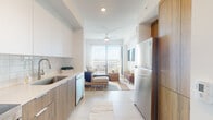 Located at 1017 Lake Ave in Lake Worth Beach, Florida, this single-story unit showcases a contemporary design defined by clean lines, a neutral palette, and an abundance of natural light throughout. The open-concept layout seamlessly connects the kitchen and living area, creating a cohesive and functional living space.

The kitchen is outfitted with two-tone cabinetry — white upper cabinets paired with warm wood-finish lower cabinets — complemented by a light quartz countertop, subway tile backsplash, stainless steel appliances including a refrigerator, dishwasher, range, and microwave, and a deep undermount sink. Large-format light tile flooring extends through the main living areas, enhancing the sense of openness.

The living area is bright and airy, anchored by a ceiling fan and floor-to-ceiling sliding glass doors that open to a private balcony offering open views of the surrounding area. A wall-mounted flat-screen TV and warm natural textures contribute to the coastal-inspired ambiance.

The bedroom features wood-look flooring, a window with natural light, and direct access to a walk-in closet. The bathroom is finished with a floating wood-tone vanity, white countertop, large mirror, and a glass-enclosed shower, maintaining the modern aesthetic found throughout the space.