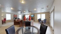 This single-floor fitness space is designed to serve as a fully functional home gym. The room's most striking feature is the bold geometric mural painted across the main wall, incorporating sharp mountain-like peaks in red, black, and neutral tones that create an energizing, athletic atmosphere. The textured popcorn ceiling adds a classic finish overhead, while two ceiling fans provide consistent air circulation throughout workouts. Natural light enters through multiple windows positioned along the walls, brightening the space without relying solely on the recessed lighting installed in the ceiling. The flooring is carpeted, offering a softer underfoot surface suited to a variety of exercise activities. Equipment visible in the room includes a treadmill, an elliptical trainer, and a cable/weight machine, providing options for both cardiovascular and strength training. A wall-mounted television is positioned for easy viewing during workouts. The layout offers ample open floor space between machines, allowing for comfortable movement and additional floor-based exercises. Overall, the room delivers a well-equipped, purpose-built workout environment with a motivating visual design.
