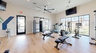 The Yardly Broadway Fitness Studio at 4550 S 67th Ave in Phoenix, AZ is a thoughtfully designed exercise room that offers a comprehensive workout experience in a bright, modern setting. The space features light hardwood-style flooring throughout, crisp white walls, and a vaulted ceiling equipped with a large ceiling fan to maintain airflow during workouts. Recessed lighting complements the abundant natural light that streams in through multiple large, black-framed windows, offering views of the surrounding outdoor landscape. The cardio zone includes treadmills, an elliptical, and a stationary bike, all featuring digital display screens. Strength training is well-represented with a multi-functional cable machine and a full rack system with a mirrored wall behind it, allowing users to monitor their form. A dedicated free weight area includes a tiered dumbbell rack stocked with a full range of weights, accompanied by adjustable benches. A wall-mounted accessory rack holds medicine balls and additional training tools. A wall-mounted TV and a water fountain add to the convenience of the space. The "Yardly Broadway" branding is prominently displayed on the accent wall, reinforcing the community identity of this well-appointed fitness amenity.