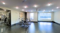 This single-floor fitness room offers a clean, functional environment designed for cardio-focused workouts. The space is anchored by a set of stationary cycling bikes arranged on a protective floor mat, providing a dedicated area for spin-style exercise. The flooring throughout is a dark hardwood-style material that adds a polished, modern aesthetic while remaining durable and easy to maintain.

Recessed lighting is evenly distributed across the ceiling, ensuring the room is well-lit at all times, while two ceiling fans promote air circulation during intense workouts. Natural light enters through multiple windows along the far wall, one of which offers a view to an outdoor balcony or corridor area, enhancing the sense of openness within the room.

The walls are finished in a neutral tone, kept largely uncluttered aside from two framed art pieces that add a touch of warmth and character. The open floor plan allows ample space for movement beyond the cycling stations, making the room adaptable for stretching or additional equipment. Overall, the space presents a bright, well-maintained fitness environment with a practical yet inviting ambiance.