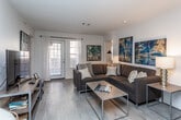 This single-floor residence offers a thoughtfully designed layout that balances comfort and functionality across its key living spaces. The living room serves as the heart of the unit, featuring light wood-style flooring, white walls, and large windows that allow natural light to fill the space. A generously sized sectional sofa anchors the room, complemented by bold blue and gold abstract artwork displayed prominently on the walls. The open sightline connects the living area to the kitchen, which features white cabinetry, gray countertops, and stainless steel appliances, including a refrigerator. An in-unit washer and dryer are conveniently located within the kitchen corridor. Two bedrooms provide restful retreats; one features sage-green walls, bold navy striped curtains, plush carpet, a ceiling fan, and space for a desk, while the second offers a more neutral palette with soft drapes and a ceiling fan. Both bathrooms are outfitted with wood-style flooring, tub-shower combinations, and white vanity cabinetry with gray countertops. One bathroom includes a mirrored sliding closet door, adding depth to the space. The overall ambiance is modern, clean, and cohesive.